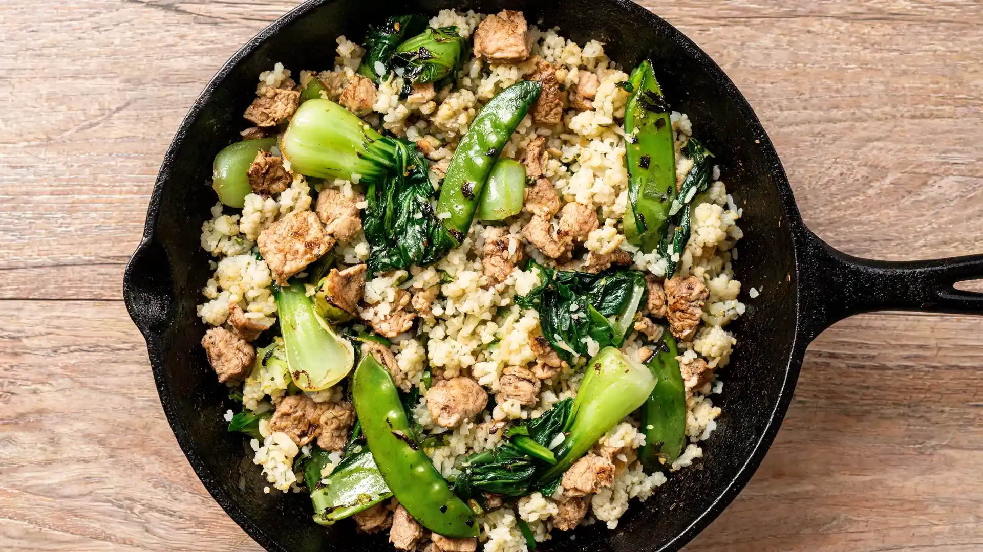 a cast iron skillet with some cauliflower rice, diced chicken, snap peas, and bok choy on a wooden table