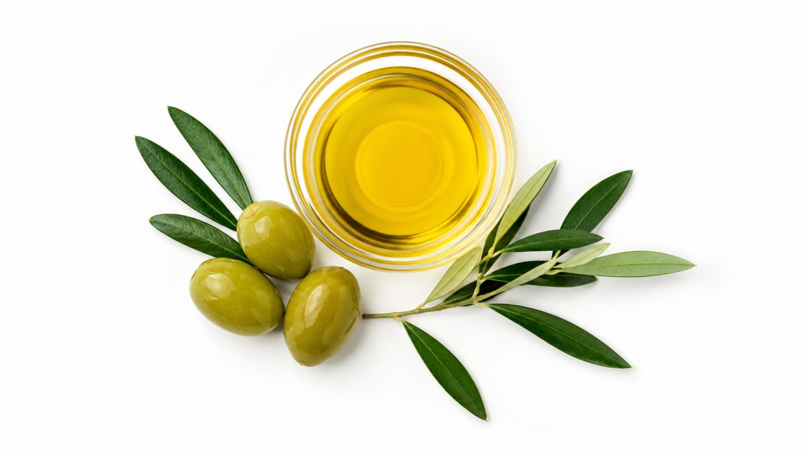 A glass bowl of golden olive oil is surrounded by three green olives and olive leaves on a white background