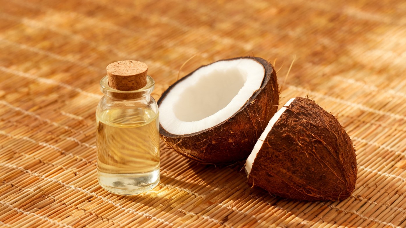 A halved coconut and a small glass bottle of oil with a cork stopper sit on a woven straw mat.