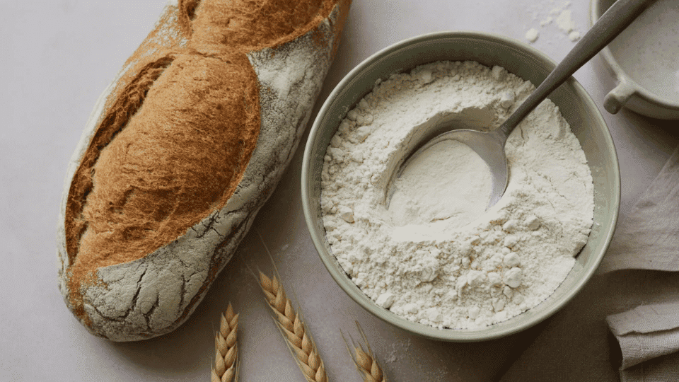 a rustic bread loaf next to a bowl of white flour with wheat stalks showing gluten ingredients