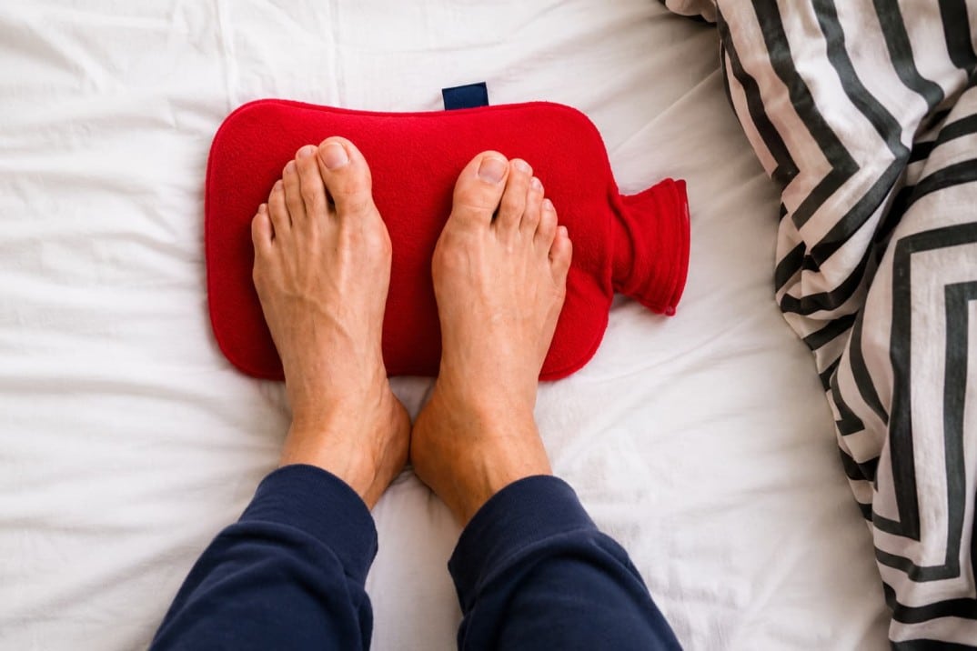 Bare feet resting on a red hot water bottle, placed on a white bed with a zigzag-patterned blanket.
