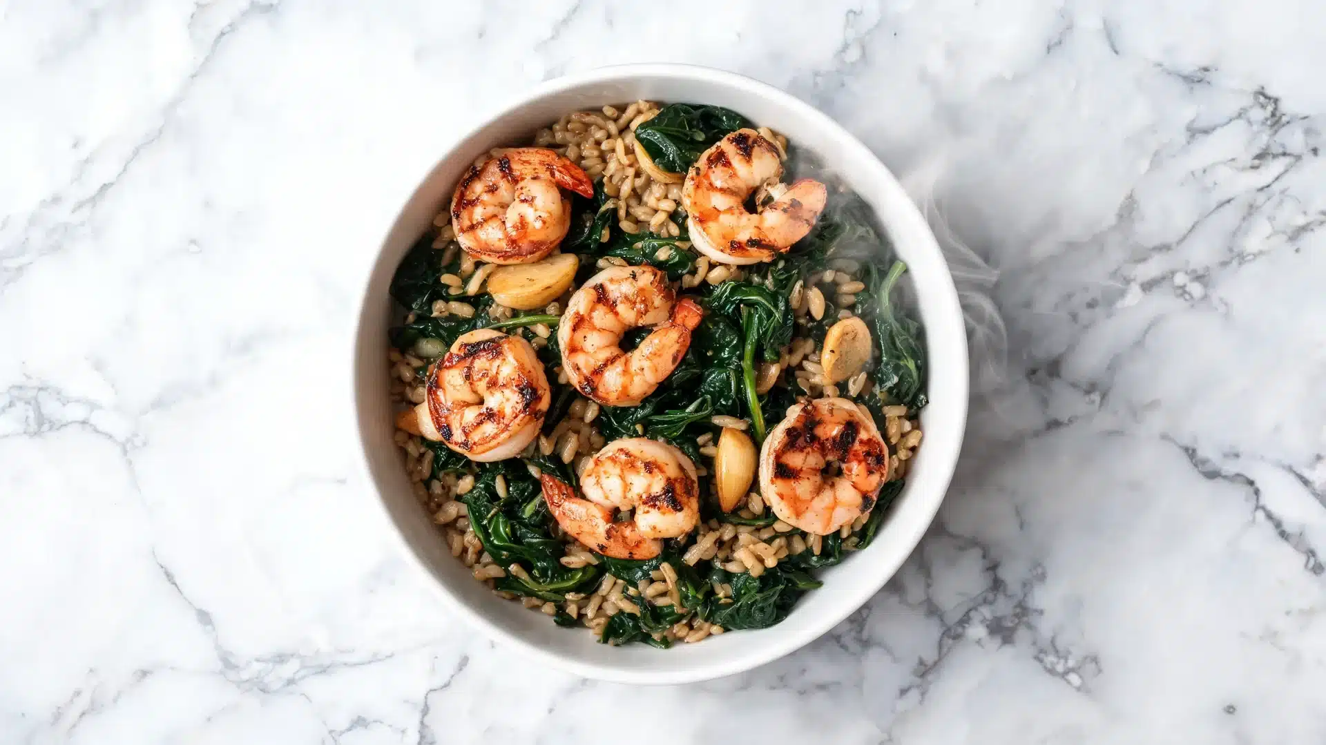bowl of grilled shrimp with brown rice, sautéed spinach, and garlic on a marble surface with visible steam