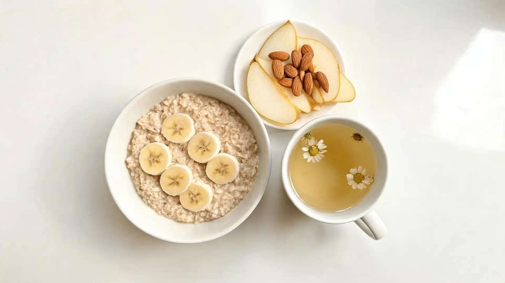 bowl of oatmeal with banana slices beside pear slices, almonds, and chamomile tea on a light-colored table surface