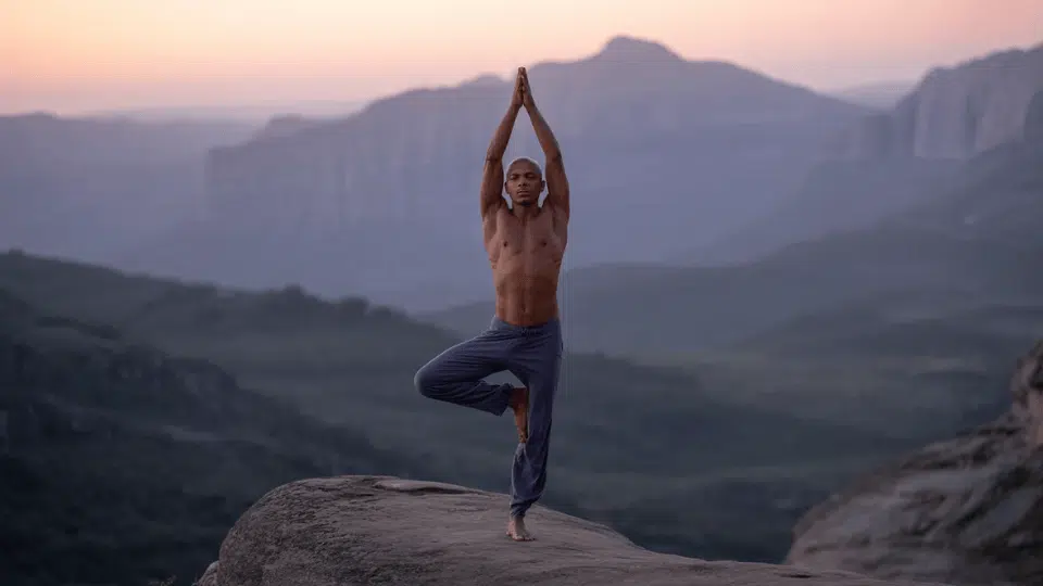 man performing tree pose on a rocky cliff edge at dusk with misty mountains in the background