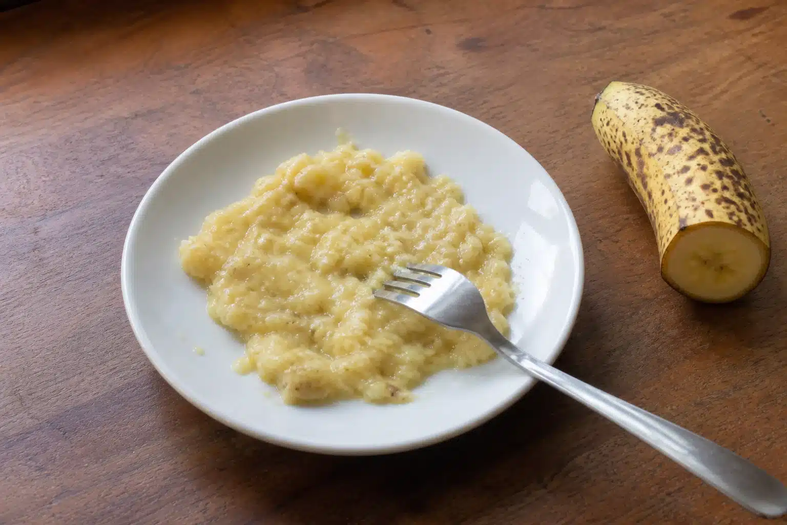 mashed ripe banana on white plate with fork, half speckled banana beside it on wooden surface in natural kitchen light