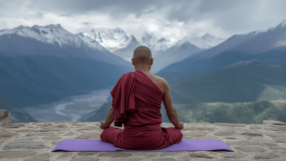 monk in red robes meditating in lotus pose on a purple mat with snow capped mountains behind