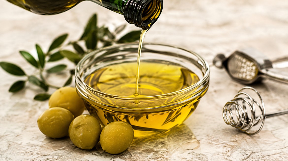 Olive oil being poured into a clear bowl surrounded by green olives and leafy branches