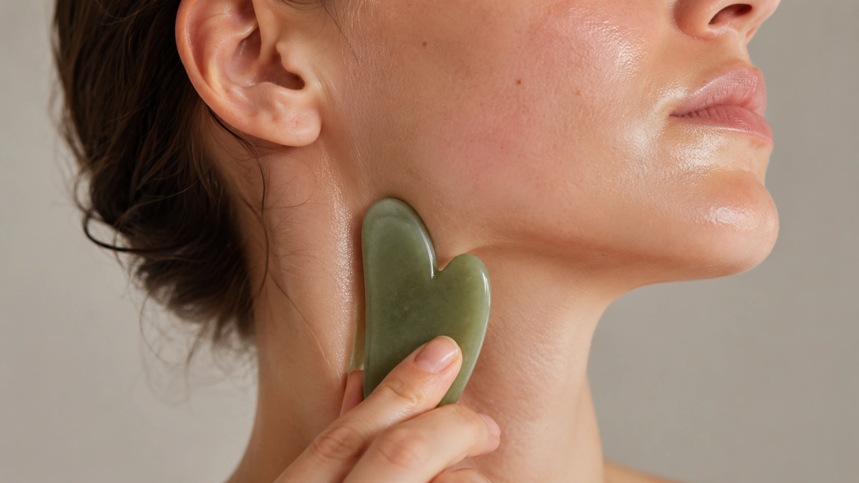 person using a green gua sha stone along the jawline and neck during a gentle skincare facial massage routine session