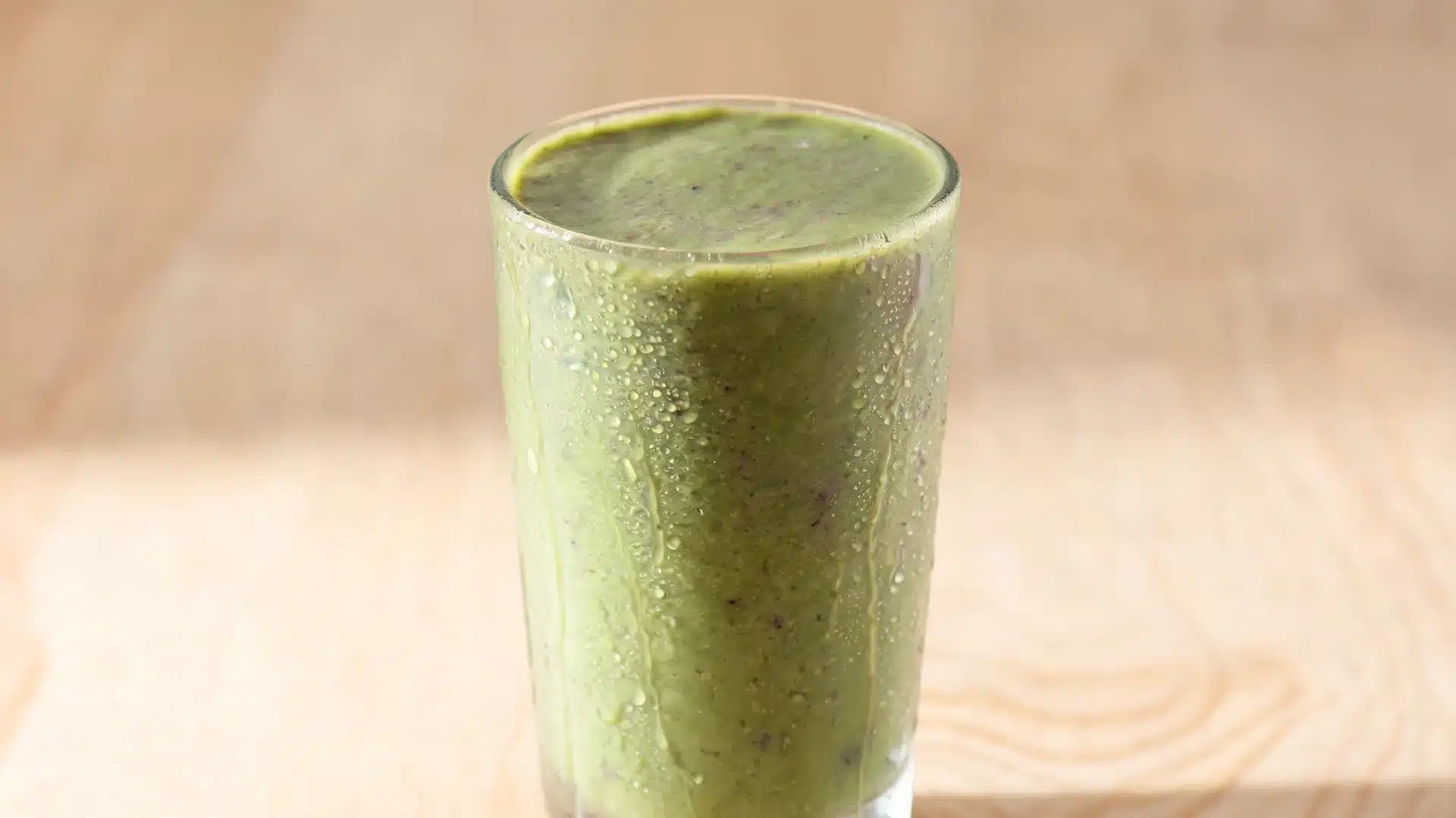 tall glass of green smoothie with condensation droplets on the surface placed on a wooden table background