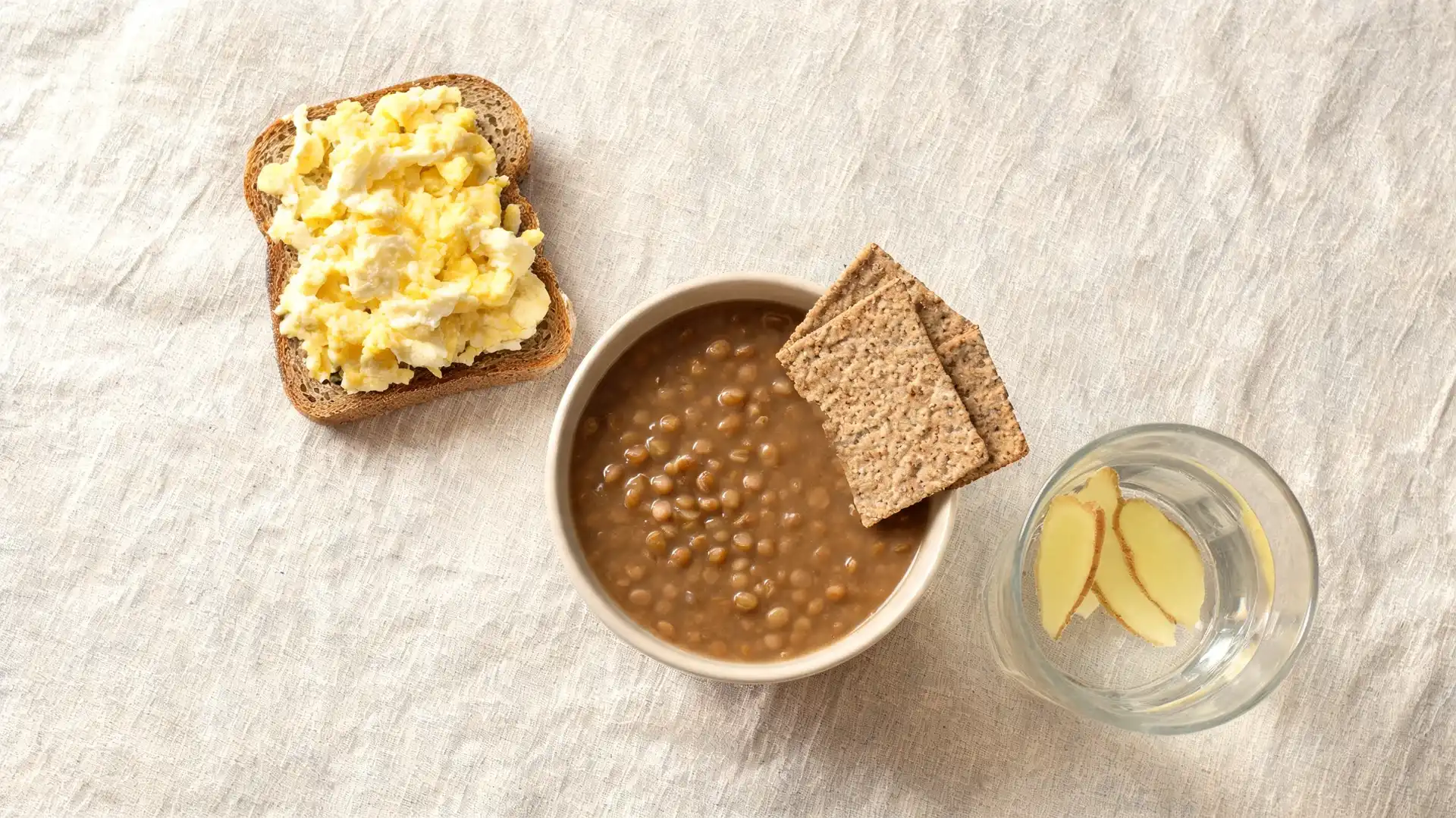 top view of lentil soup with crackers, scrambled eggs on toast, and ginger water served on a light fabric surface