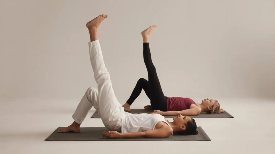 two people practicing supine leg raise yoga poses on mats against a neutral background