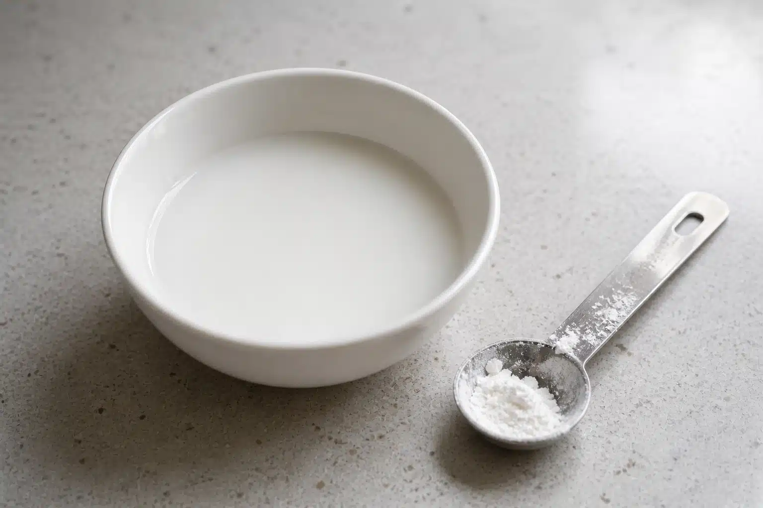 white bowl with smooth cornstarch and water mixture on light countertop, metal measuring spoon holding powder beside