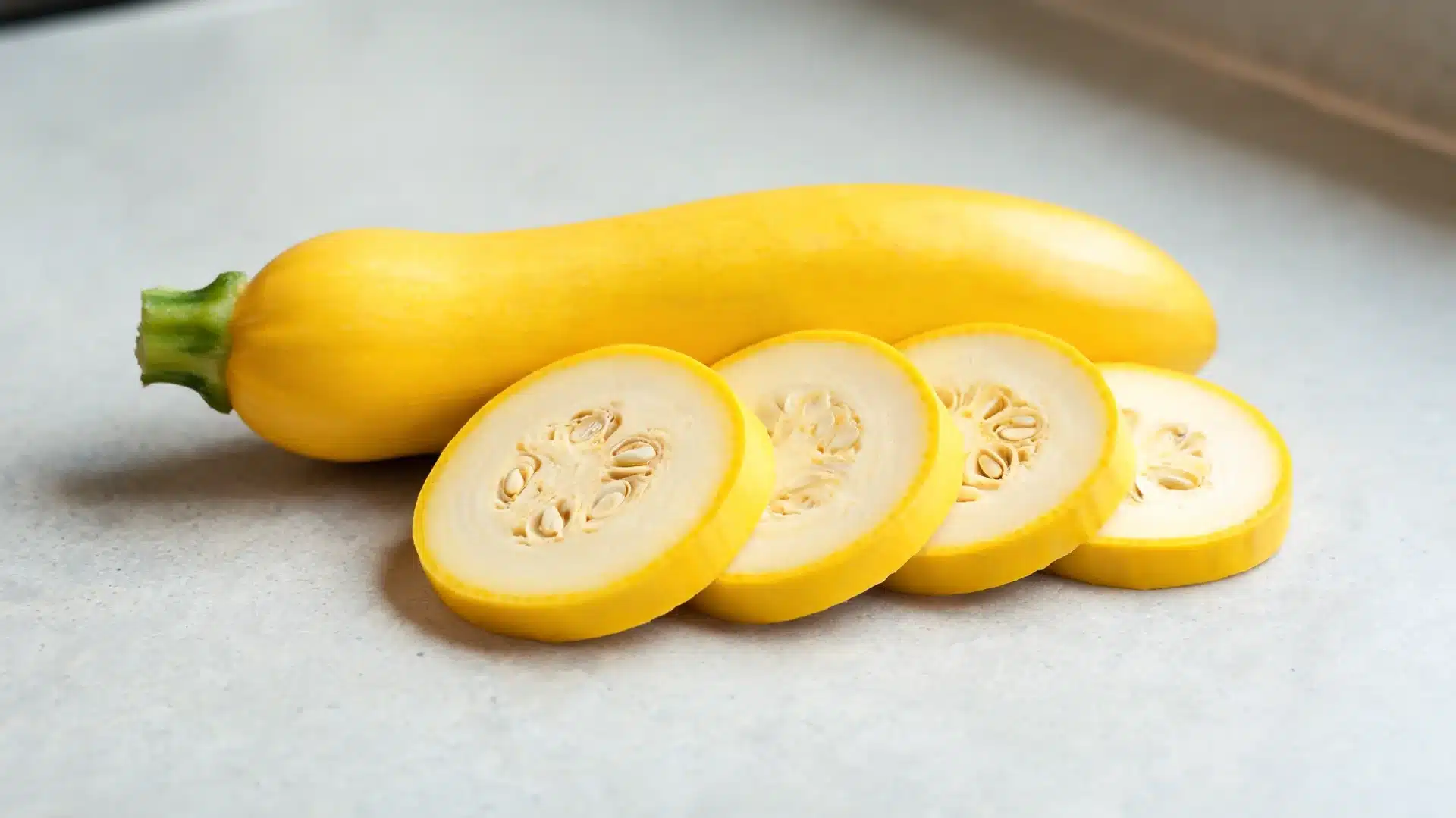 whole yellow squash with smooth skin and several round slices showing pale interior and seeds, arranged on light surface