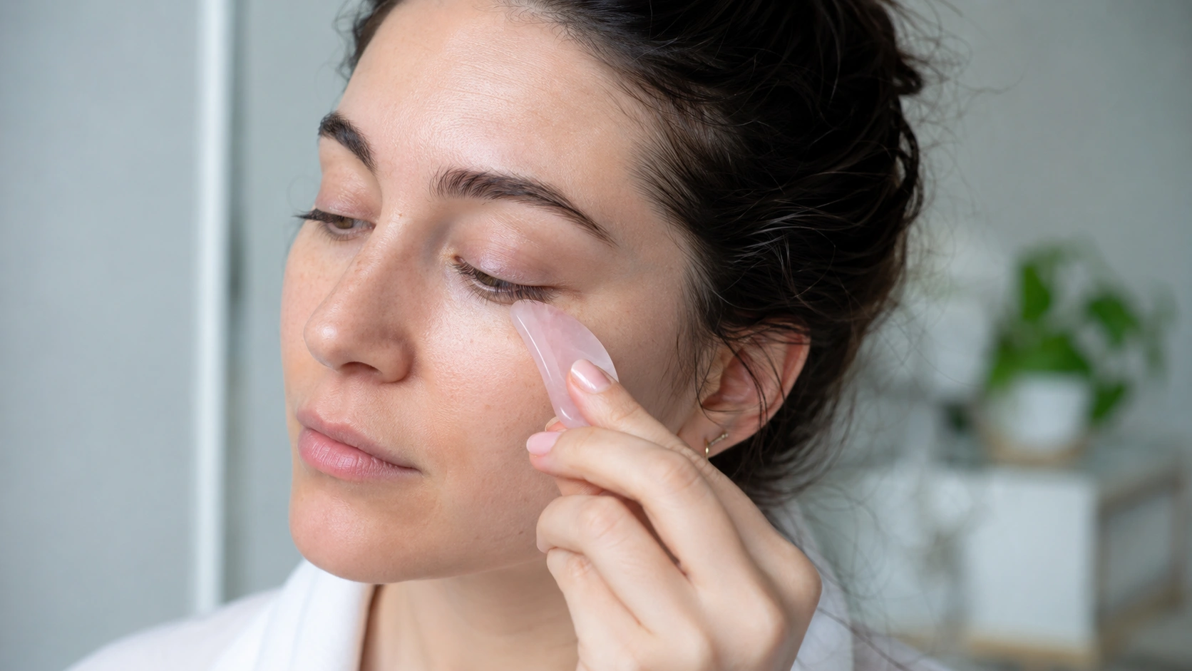 woman gently uses a rose quartz gua sha tool under her eye during a bright facial skincare routine