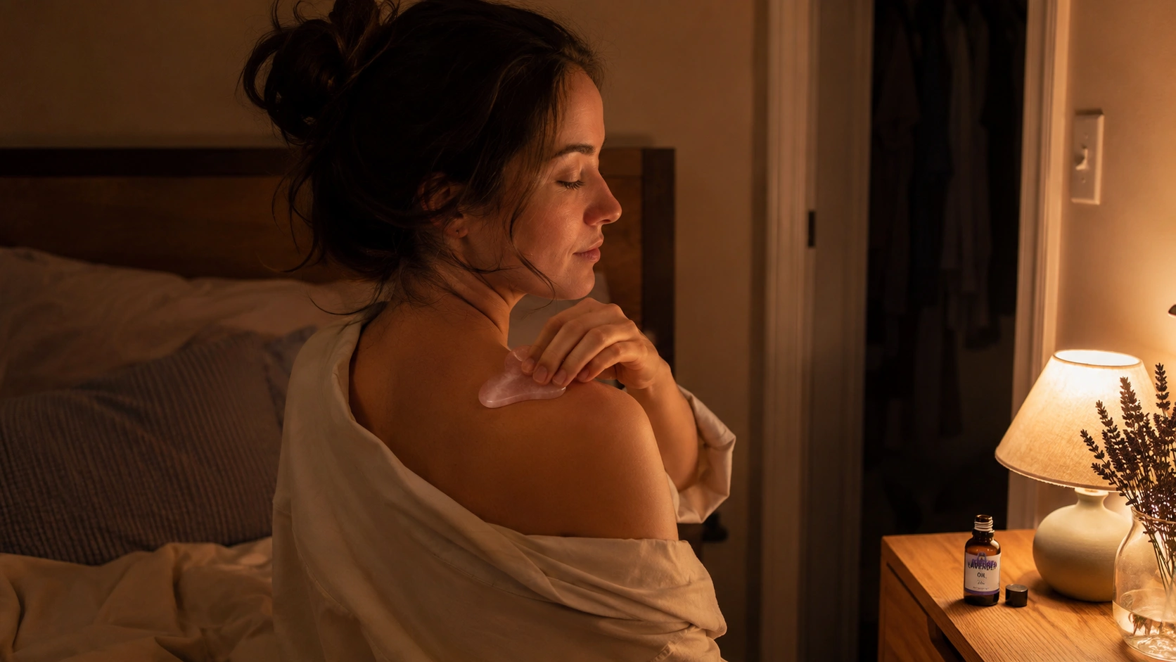 woman uses a rose quartz gua sha tool on her shoulder during a warm nighttime self-care routine in bed