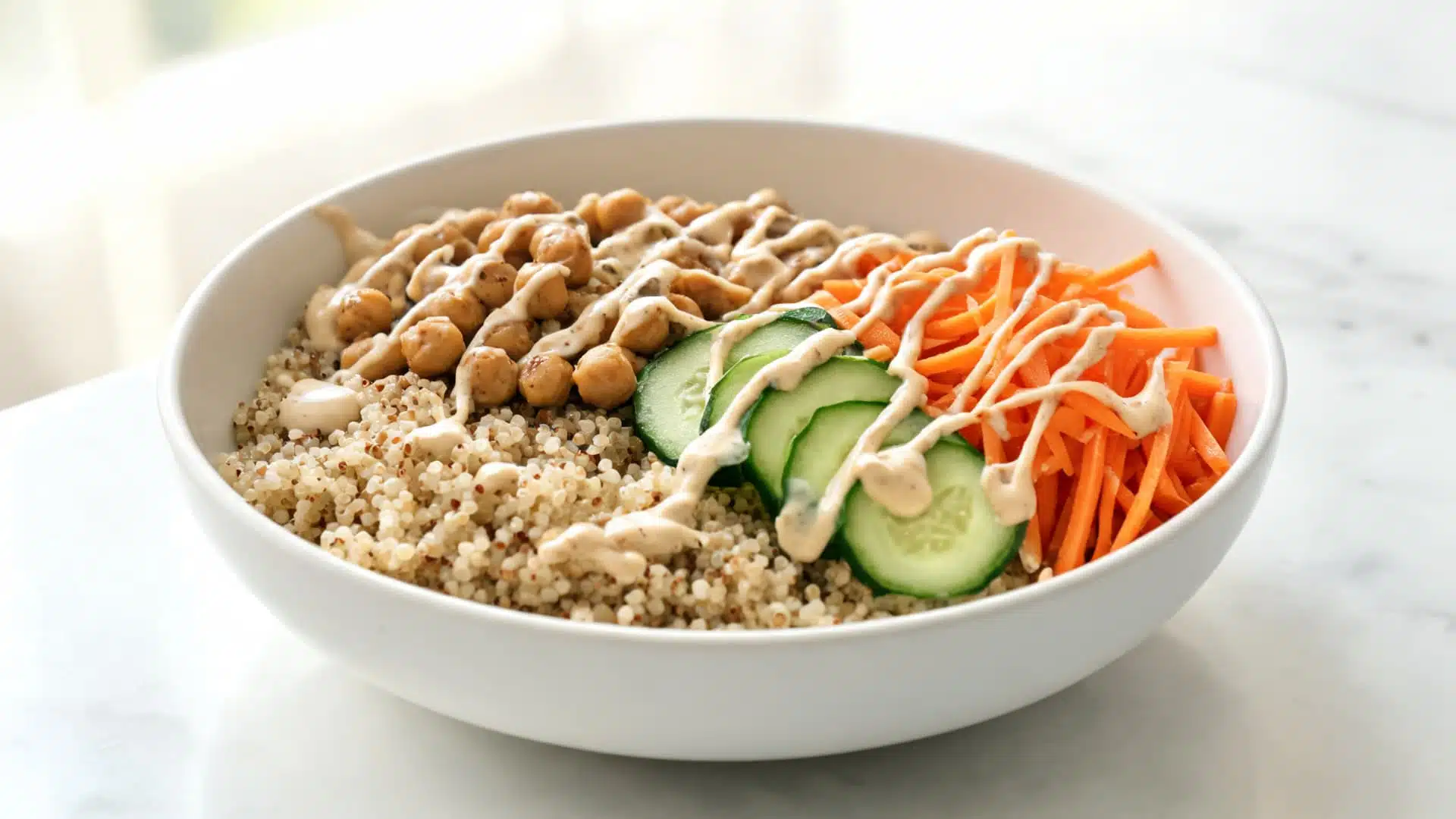 bowl of quinoa topped with chickpeas cucumber shredded carrot and creamy dressing served on marble surface in soft natural light