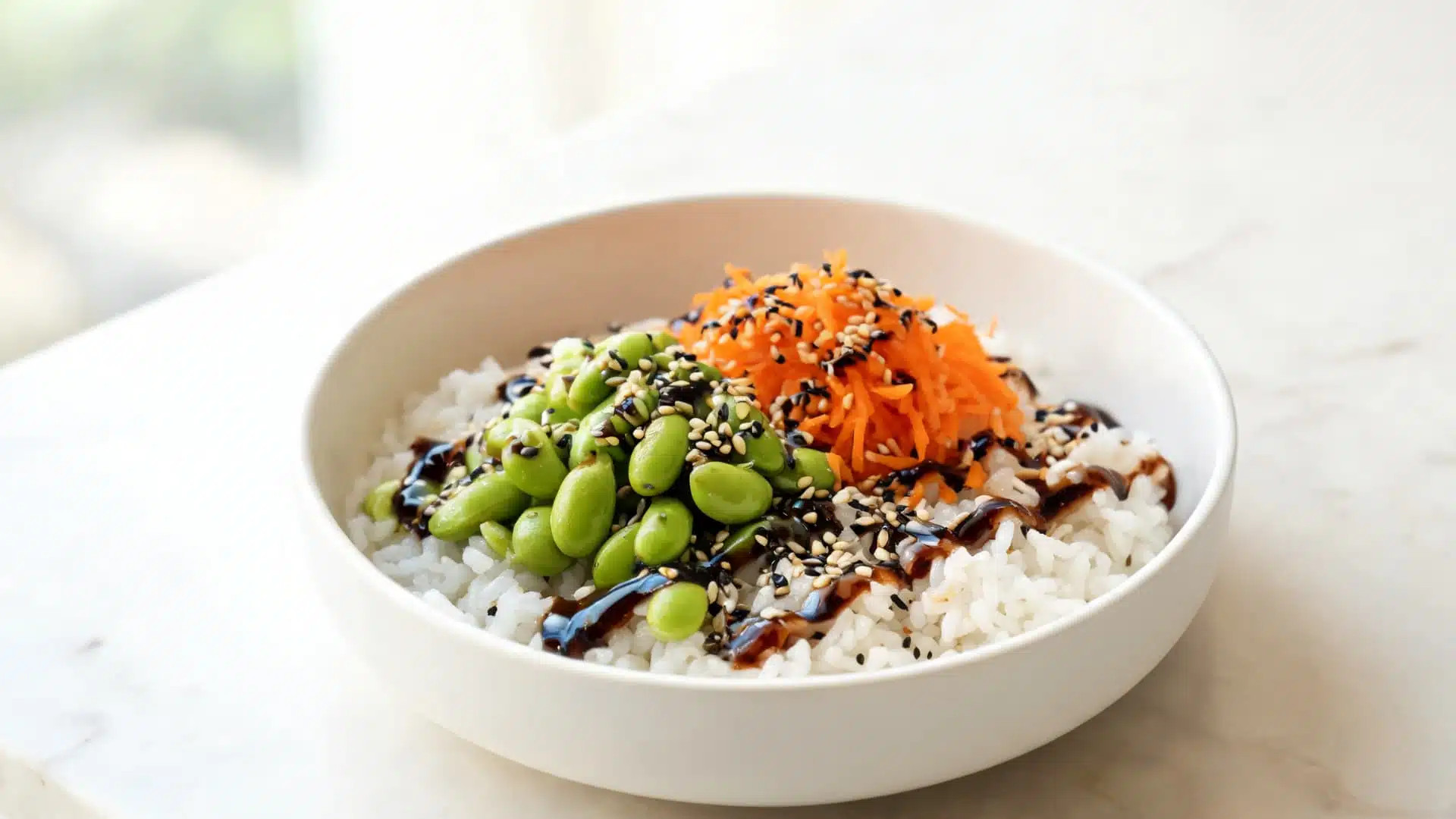 bowl of rice topped with edamame shredded carrot sesame seeds and dark sauce on marble countertop
