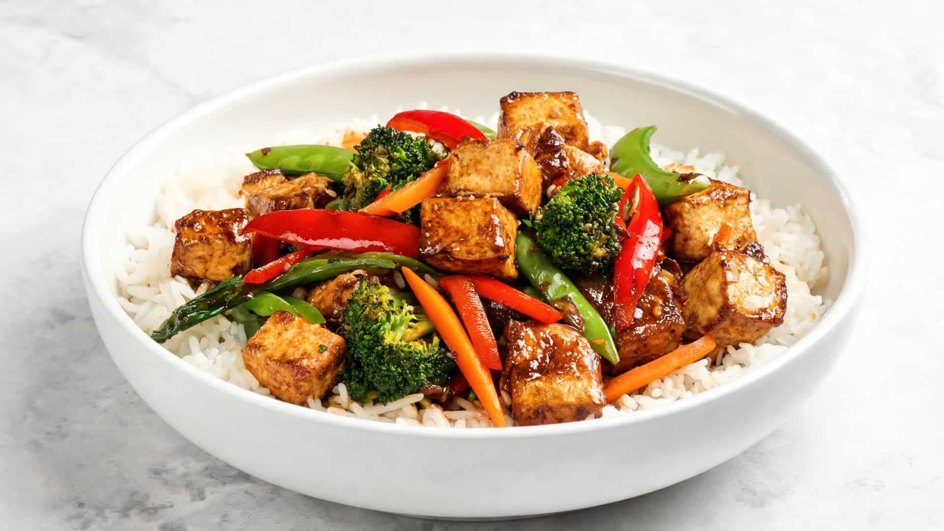 bowl of rice topped with glazed tofu broccoli and bell peppers in savory sauce on marble countertop bright natural light