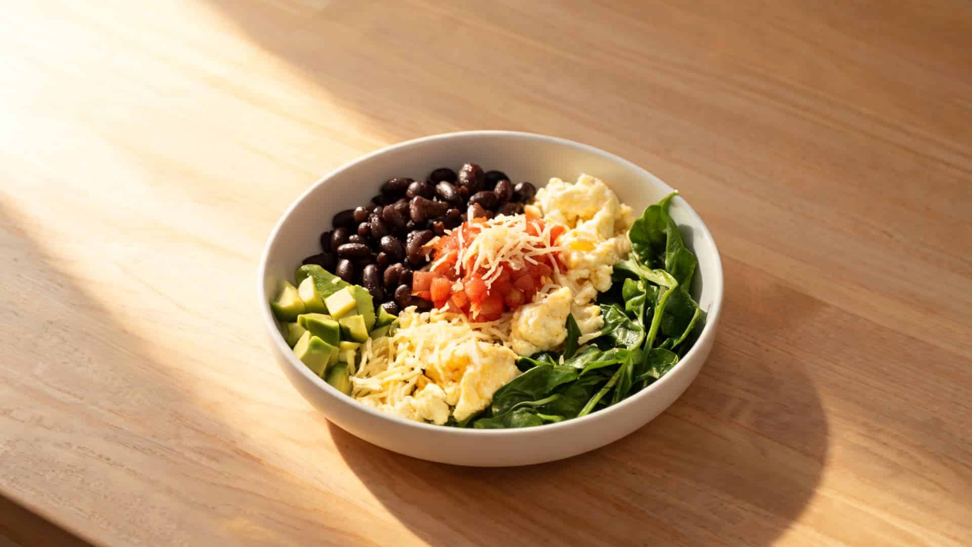 breakfast bowl with scrambled eggs, spinach, avocado, black beans, salsa and cheese on a sunny wooden table (1)