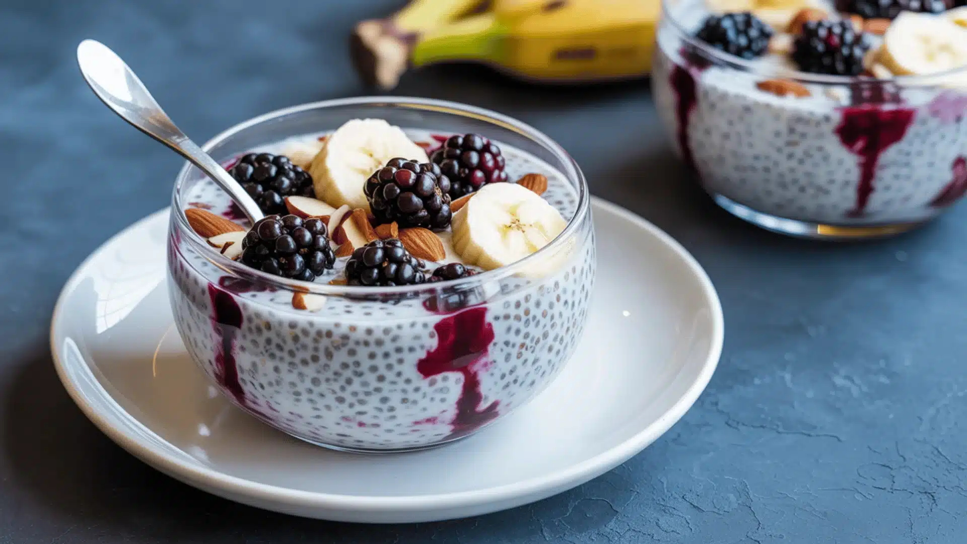 chia seed pudding in a clear glass bowl on a white ceramic saucer the pudding contains dark blue-black blackberries