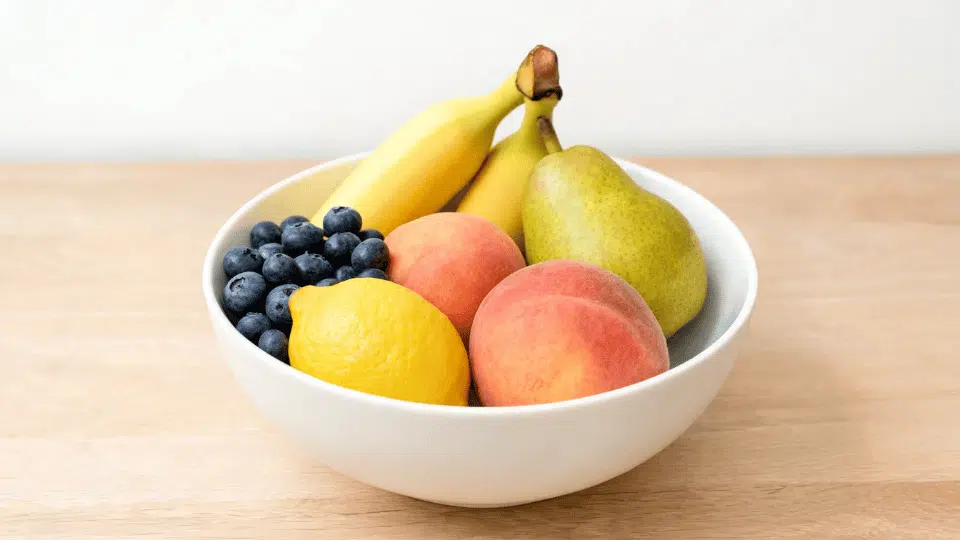 fruit bowl with bananas, blueberries, lemon, peaches, and pear on a wooden table