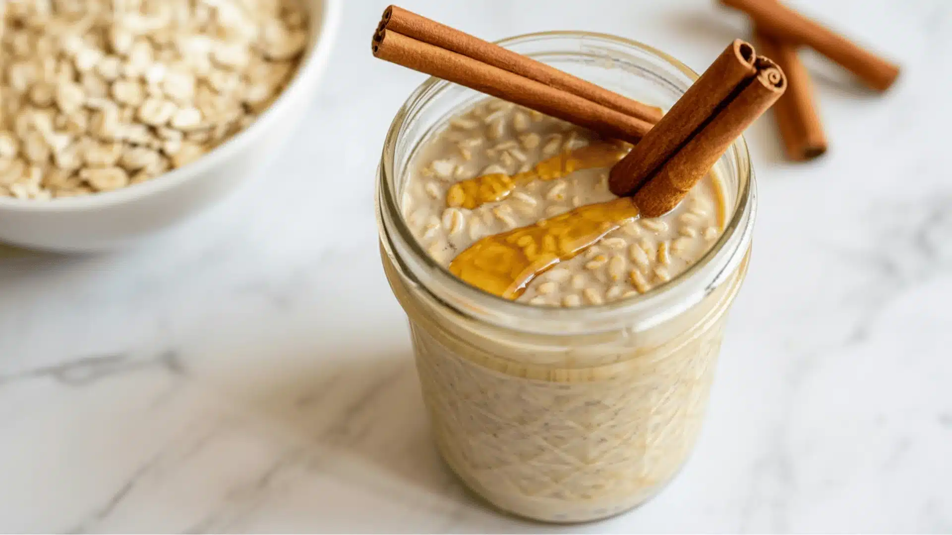 glass mason jar on a white marble surface the oats are topped with two whole cinnamon sticks placed diagonally across the to