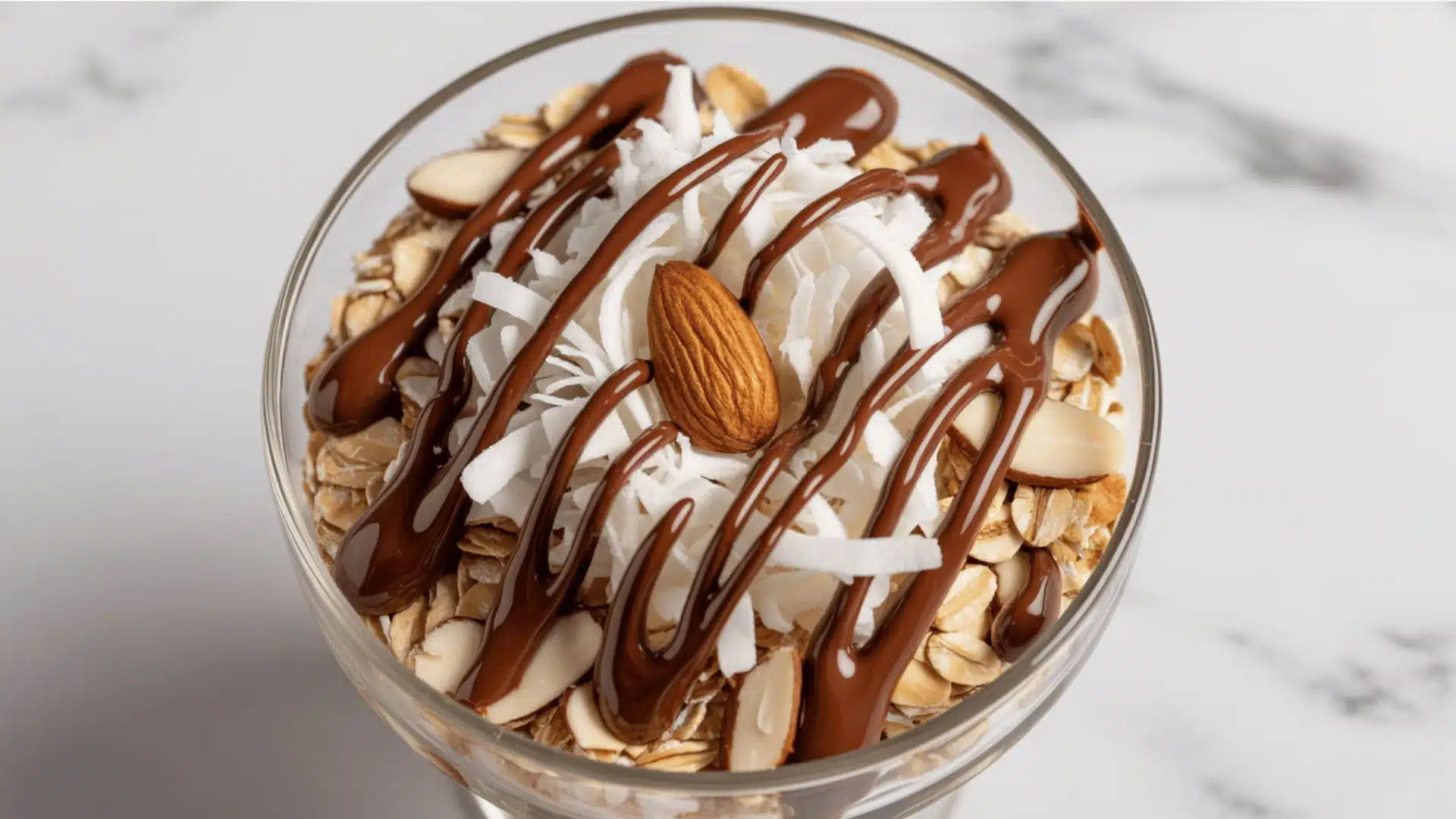 gourmet dessert topping mixture in a clear glass bowl consists of white shredded coconut, oats, sliced almonds, and dark chocolate