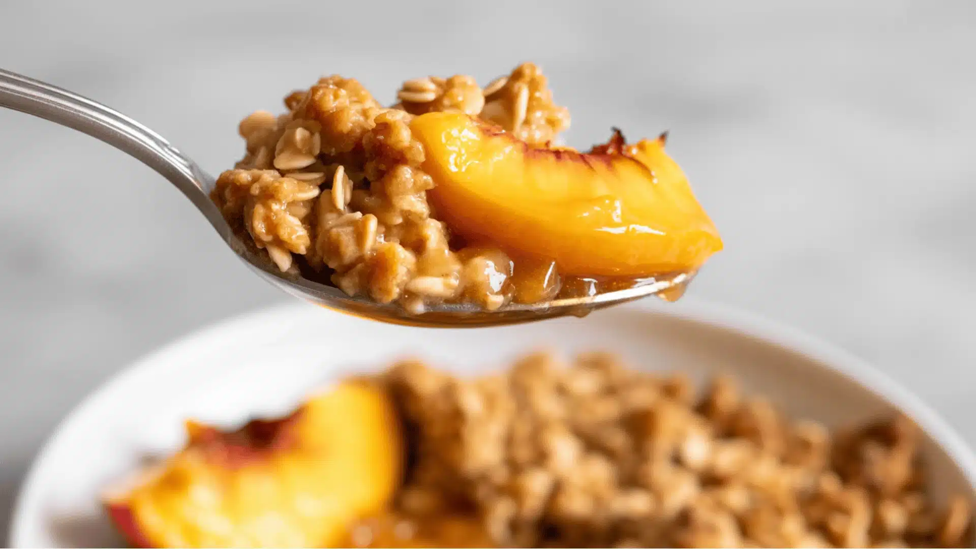 light gray background the spoon contains golden-brown oatmeal topping with chunks of ripe, caramelized peach slices