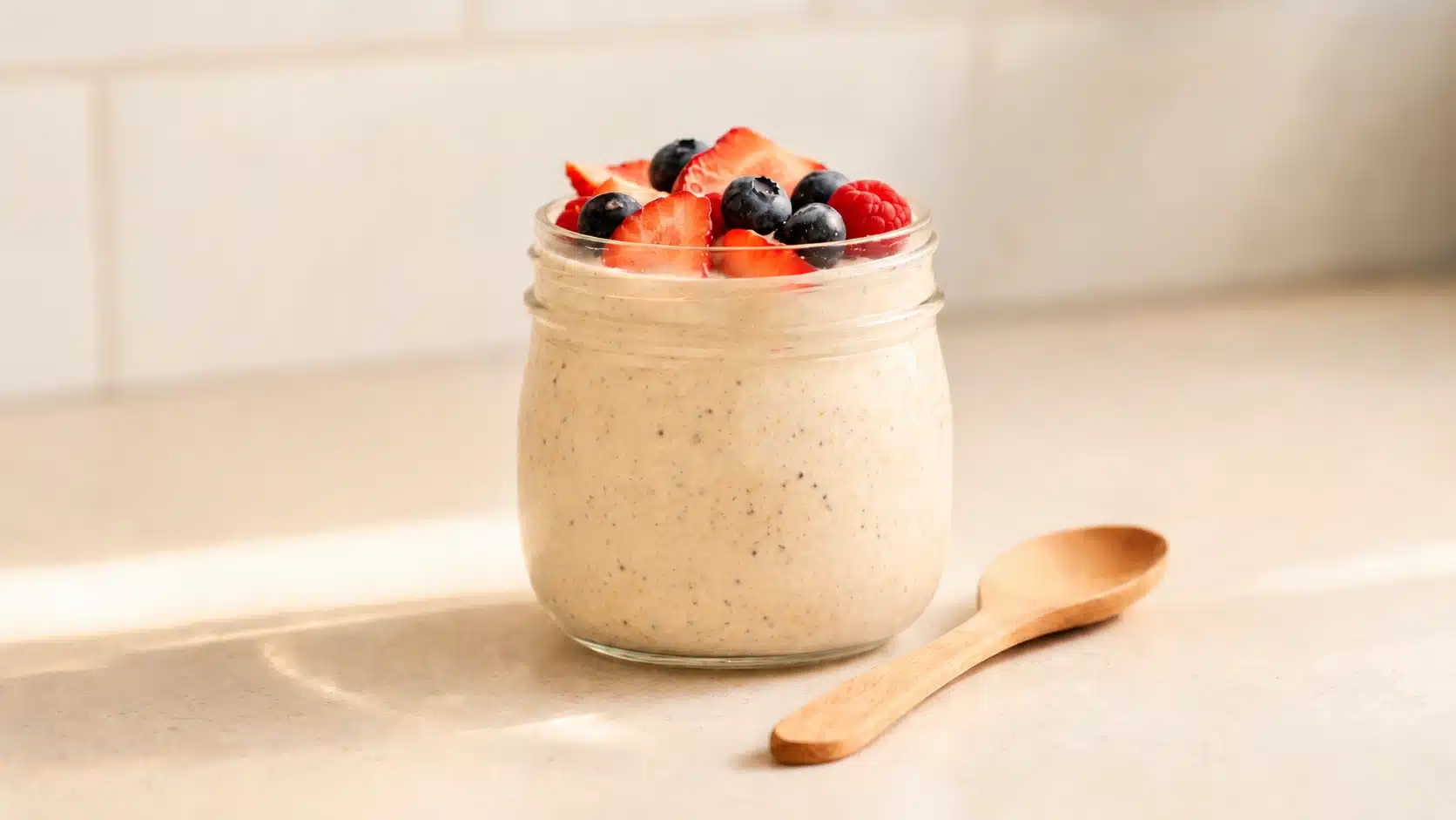 overnight oats in a glass jar topped with strawberries, blueberries and raspberries beside a wooden spoon