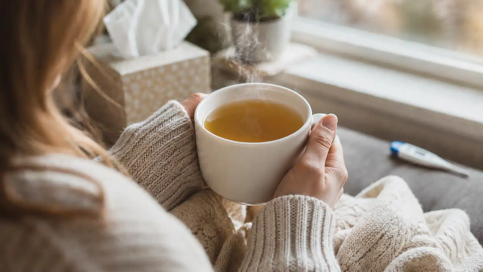 person holding warm medicine ball tea near a window during a quiet sick day at home