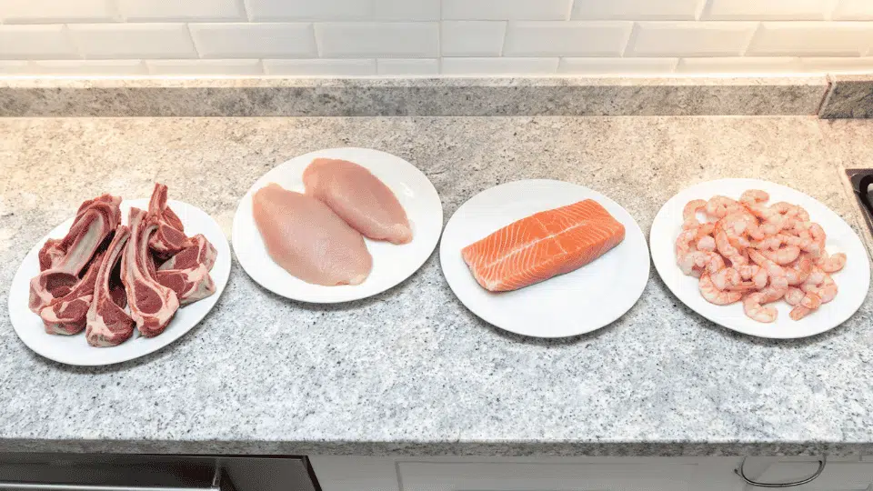 raw lamb chops, chicken, salmon, and shrimp served on white plates on a kitchen counter