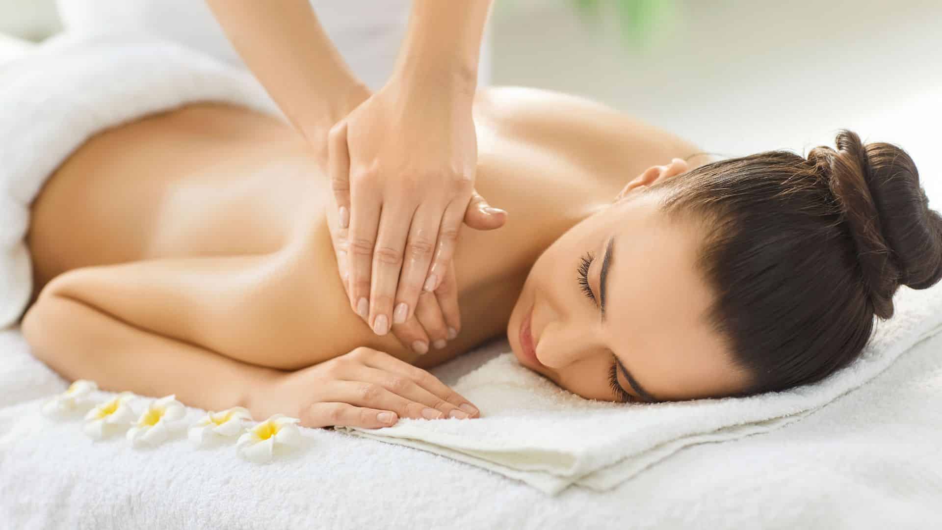 serene woman with her hair in a bun lies on a massage table, smiling with closed eyes while a therapist massages her upper back and shoulder
