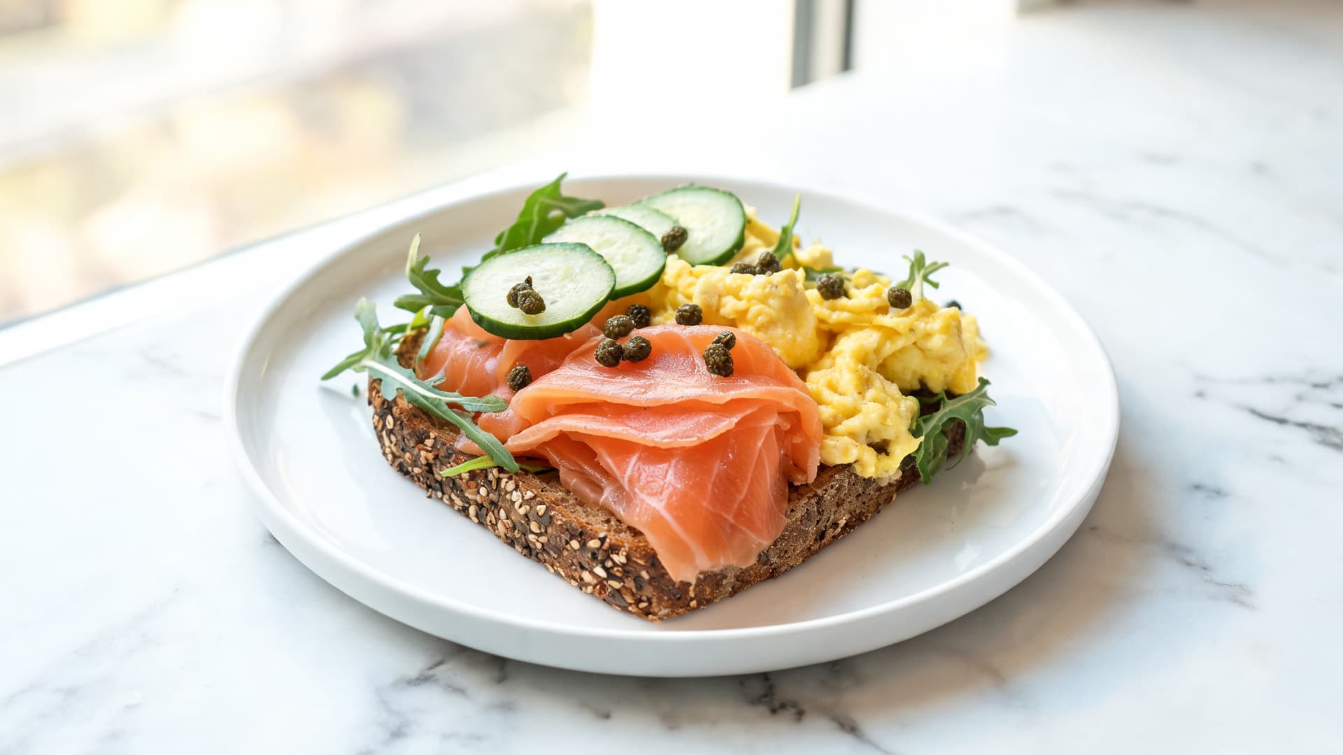 smoked salmon toast with scrambled eggs, cucumber, arugula, and capers on a marble counter