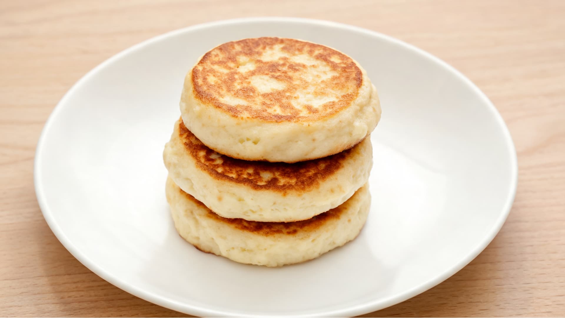 three golden cottage cheese pancakes stacked on a white plate over a light wooden table