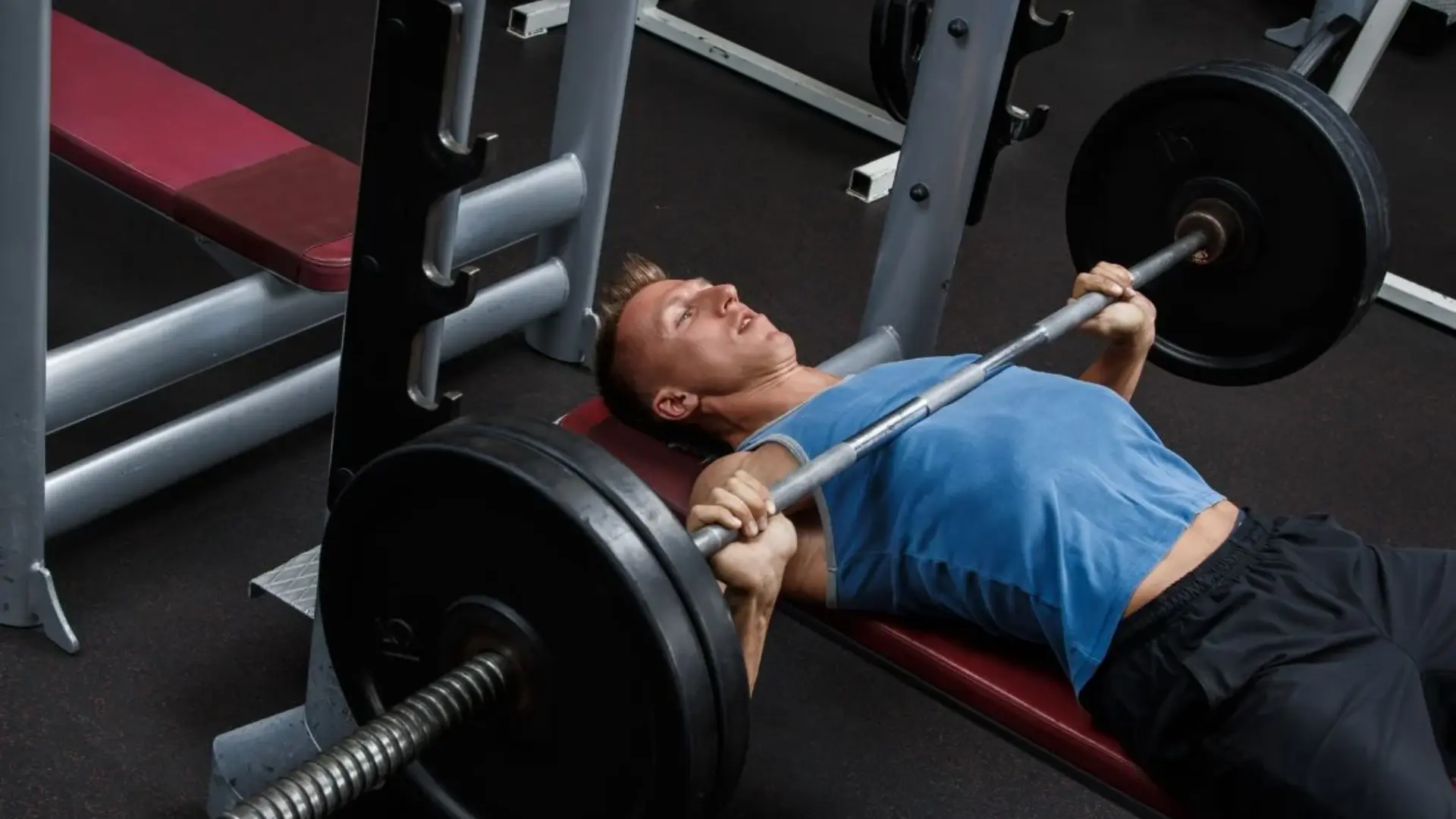 a man performs a heavy barbell bench press in a home gym, gripping the bar with both hands as he lifts it off the rack, showing the intense effort of anaerobic exercise.