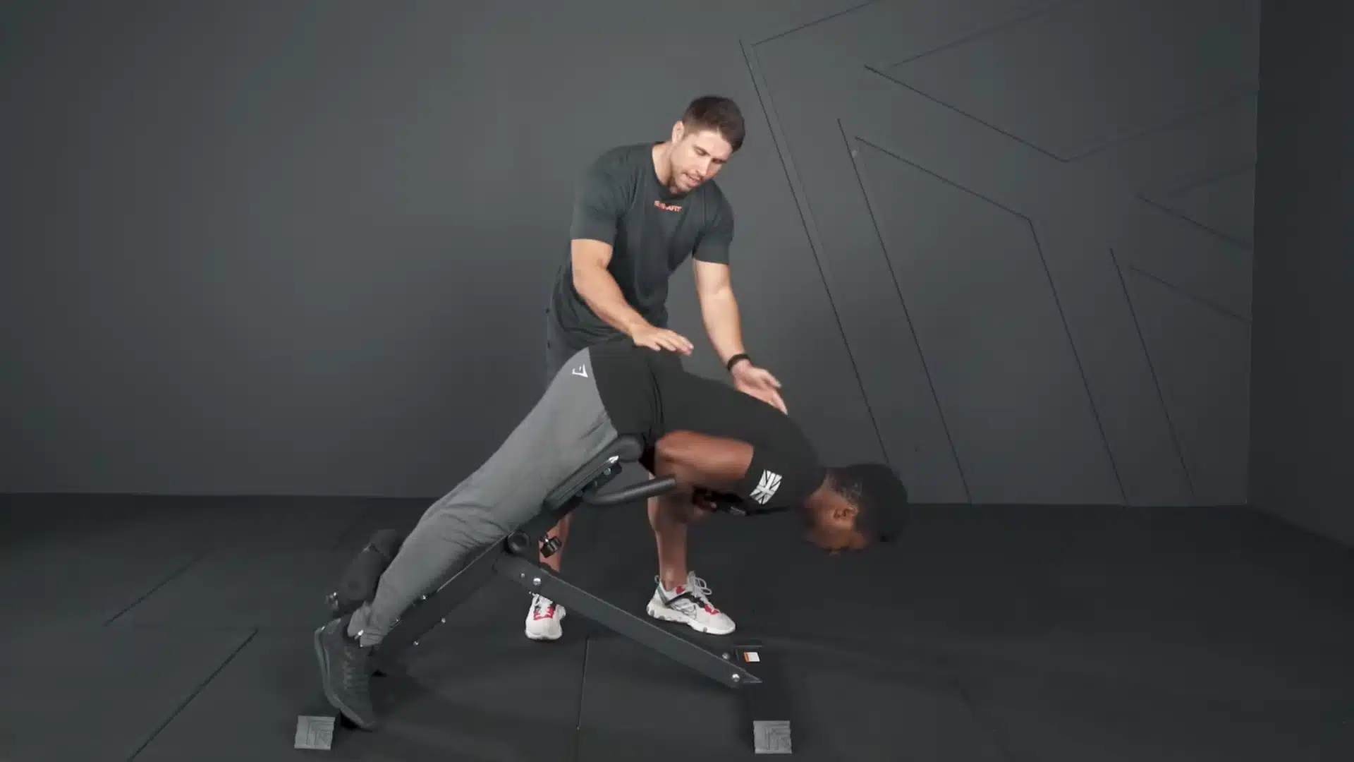 trainer guides an athlete through the lowered position of a hyperextension on an angled bench