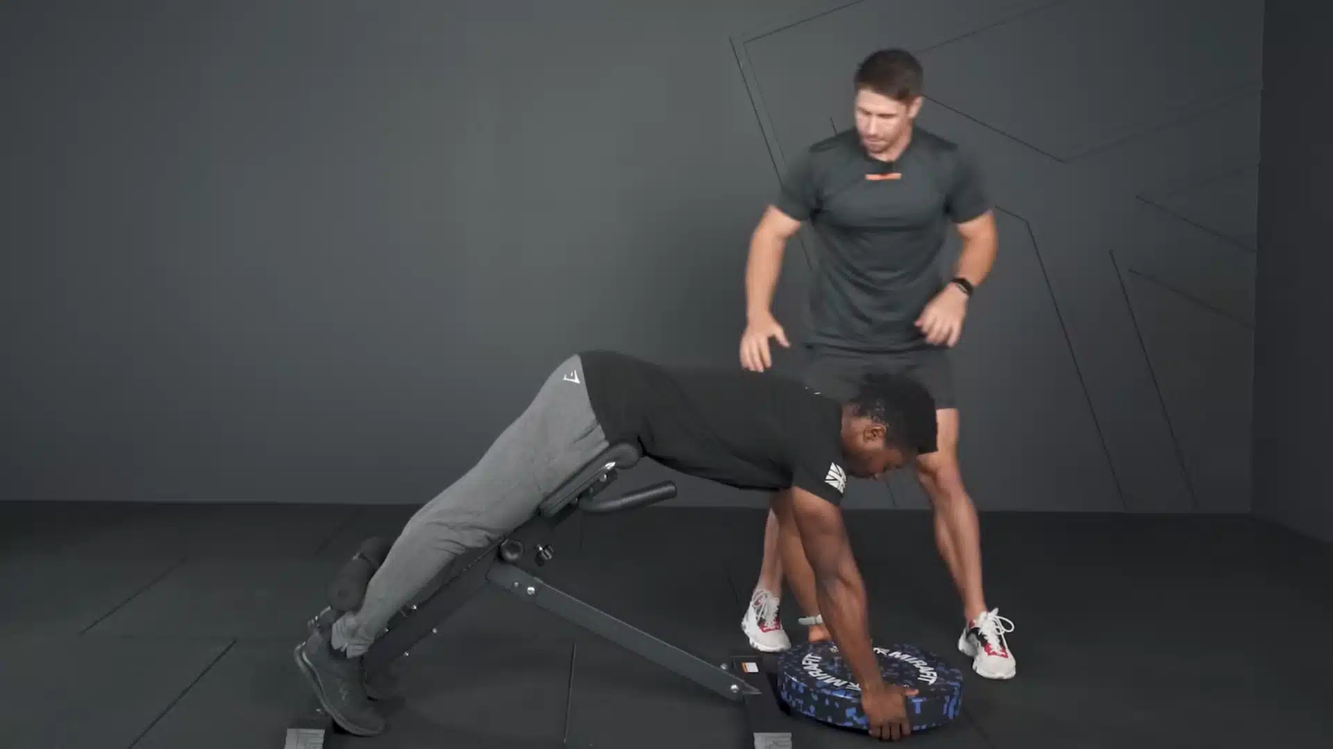 athlete reaches from a hyperextension bench to lift a weight plate while a trainer supervises